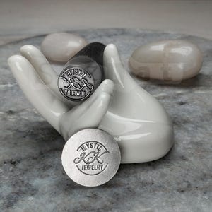 May include: A white ceramic hand holds two metal stamps with the text "Mystic KK Jewelry". One stamp is round, the other is cylindrical. The stamps are silver-colored. Two smooth, white stones are in the background.