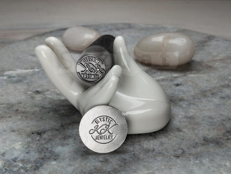 May include: A white ceramic hand holds two metal stamps with the text "Mystic KK Jewelry". One stamp is round, the other is cylindrical. The stamps are silver-colored. Two smooth, white stones are in the background.