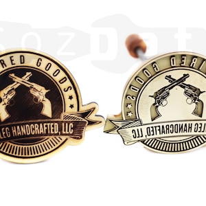 May include: Two wooden branding irons with a circular design featuring crossed revolvers and the text "INSPIRED GOODS" and "HOGLEG HANDCRAFTED, LLC". One iron is light wood, the other is metallic.