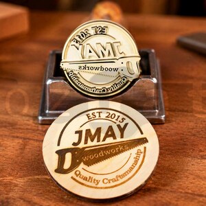 May include: Two custom wood stamps with circular designs and the text "JMAY woodworks" and "AMC". The stamps feature a saw graphic and the words "Quality Craftsmanship" and "EST 2015". One stamp is in a metal holder. The background is a wooden surface.