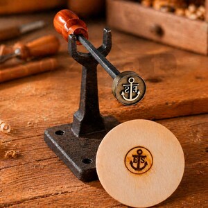 May include: A woodworking tool with a dark metal base and handle, topped with a wooden handle. The tool has an anchor emblem. A wooden disc with the same anchor emblem is next to the tool. The background includes other woodworking tools.
