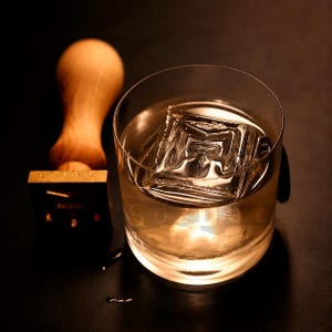 Puede incluir: Un vaso transparente con líquido ámbar y un gran cubito de hielo cuadrado. Un sello de madera con base dorada en el fondo. El cubito de hielo tiene un diseño. El vaso está sobre una superficie oscura.