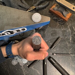 May include: A collection of metalworking tools and components, including a stamp with a circular design, pliers, and a small anvil. The stamp is held in a hand, with several other tools scattered around.