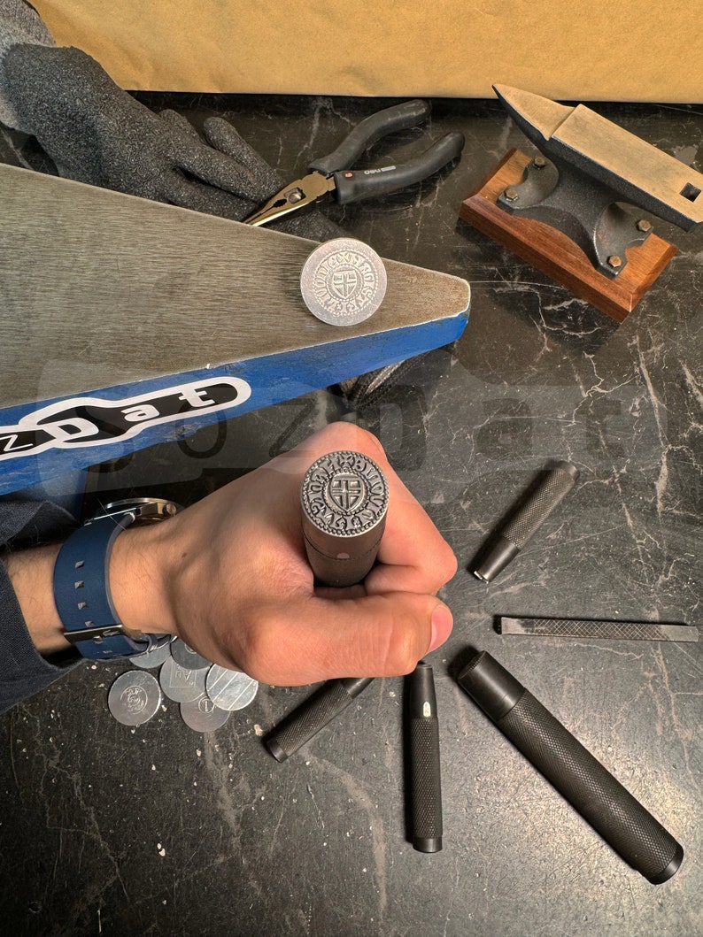 May include: A collection of metalworking tools and components, including a stamp with a circular design, pliers, and a small anvil. The stamp is held in a hand, with several other tools scattered around.
