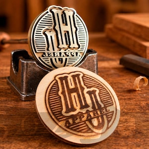 May include: Two wooden stamps with the text "H Apparel U" and a metal stamp holder. The stamps are circular with a detailed design. The background includes woodworking tools and wooden blocks, suggesting a craft or design theme.