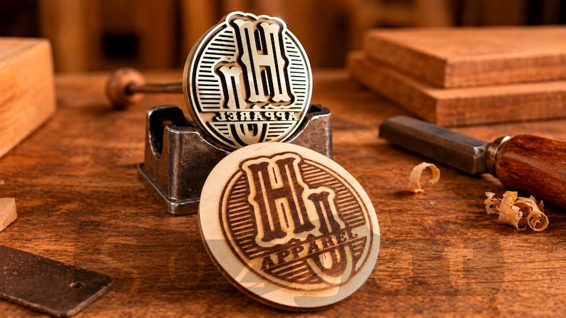 May include: Two wooden stamps with the text "H Apparel U" and a metal stamp holder. The stamps are circular with a detailed design. The background includes woodworking tools and wooden blocks, suggesting a craft or design theme.