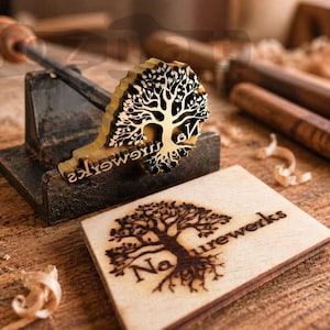 May include: A wooden stamp with a tree design and the word "Naturewerks" is displayed on a wooden surface. The stamp is mounted on a metal base with a wooden handle. A wooden block with the same tree design is also present.