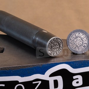 May include: A metal stamp set with a handle and a circular die. The die features a detailed, symmetrical design with a cross at the center. The handle is textured for grip, and the set rests on a metal surface.