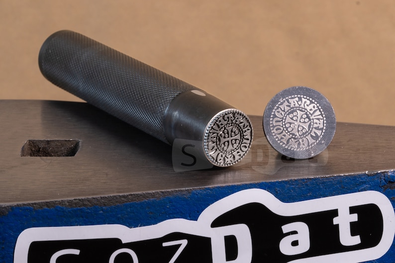 May include: A metal stamp set with a handle and a circular die. The die features a detailed, symmetrical design with a cross at the center. The handle is textured for grip, and the set rests on a metal surface.