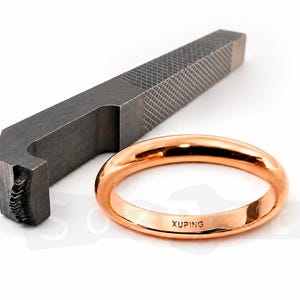May include: A rose gold ring with the word "XUPING" engraved on it, next to a dark gray metal tool with a textured surface. The ring is smooth and shiny, reflecting the light. The tool has a rectangular shape with a patterned top.
