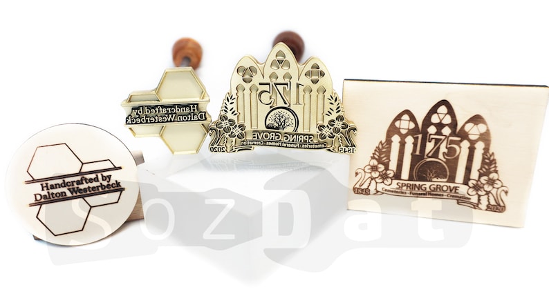 May include: A collection of custom stamps with wooden handles. The stamps feature intricate designs, including text that reads "Handcrafted by Dalton Westerbeck" and "Spring Grove 175". The stamps are in various shapes and sizes.