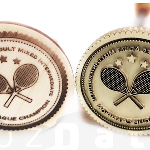 May include: Two tennis champion medals. One is made of wood with a dark brown engraved design, the other is gold-colored. Both feature crossed tennis rackets, stars, and text that reads "Wong-Holmes Adult Mixed Intermediate League Champion."