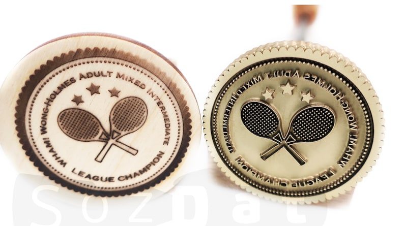 May include: Two tennis champion medals. One is made of wood with a dark brown engraved design, the other is gold-colored. Both feature crossed tennis rackets, stars, and text that reads "Wong-Holmes Adult Mixed Intermediate League Champion."