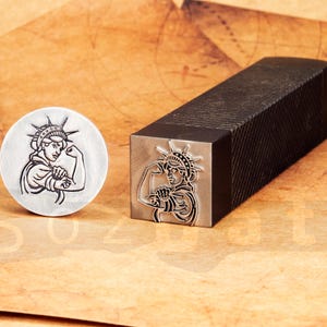 Könnte beinhalten: Ein runder Metallstempel und ein rechteckiger Metallstempel mit schwarzem Griff. Beide Stempel zeigen eine Grafik der Freiheitsstatue, die ihren Bizeps anspannt. Die Stempel befinden sich auf einer braunen Oberfläche.
