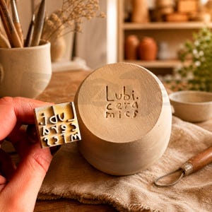 Könnte beinhalten: Ein hellbrauner Keramikstempel und eine passende zylindrische Basis. Der Stempel wird in der Hand gehalten und hat eine quadratische Form mit Zahlen und Buchstaben. Die Basis trägt den eingravierten Text "Lubi. ceramics".