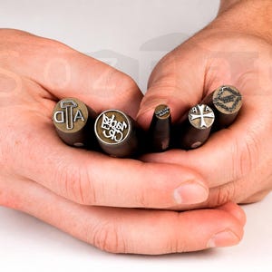 May include: A collection of metal stamps with wooden handles, each featuring a different design. The stamps include letters, symbols, and a cross. The stamps are held in open hands against a white background.