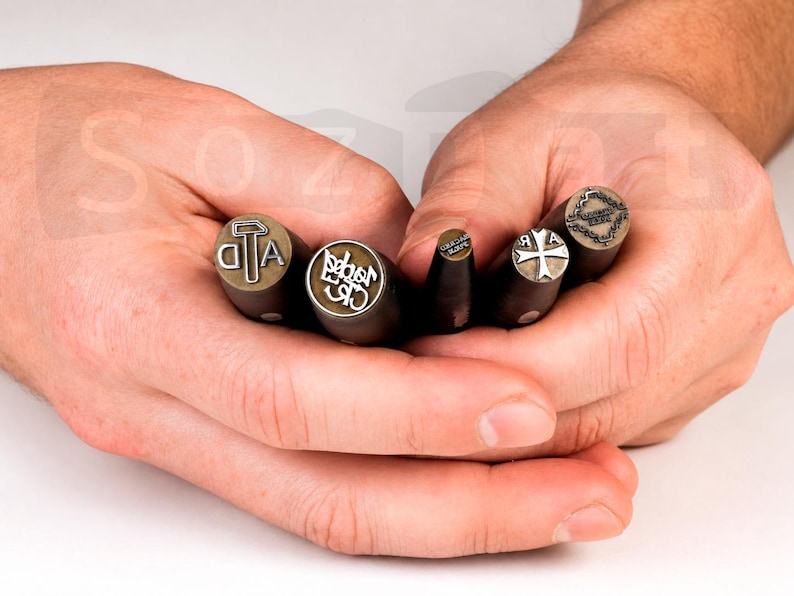 May include: A collection of metal stamps with wooden handles, each featuring a different design. The stamps include letters, symbols, and a cross. The stamps are held in open hands against a white background.
