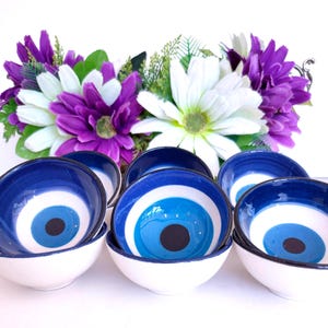 May include: A set of small ceramic bowls featuring a concentric blue, white, and black eye pattern on the interior. The bowls have a smooth white exterior and are arranged in front of a cluster of artificial purple and white daisy flowers.