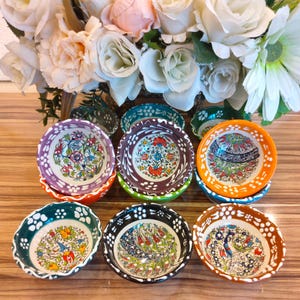 Può includere: Set di otto piccole ciotole in ceramica con motivi floreali colorati. Le ciotole hanno un bordo ondulato e sono disposte in cerchio su una superficie di legno.