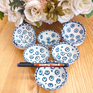 Puede incluir: Seis cuencos pequeños de cerámica blanca con un diseño floral azul y bordes festoneados. Los cuencos están dispuestos sobre una superficie de madera, con un rotulador negro que dice "edding 140 S" en el centro. Flores blancas en el fondo.