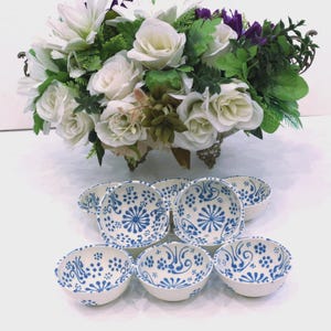 May include: A collection of seven small white ceramic bowls featuring a hand-painted blue floral pattern with intricate swirls and dots. The bowls are arranged in a cluster below a decorative centrepiece of white roses and green foliage.