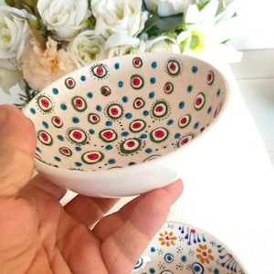 May include: Two white ceramic bowls with blue, green, and red eye designs. The bowls are hand-painted with a white background and feature a variety of eye patterns.