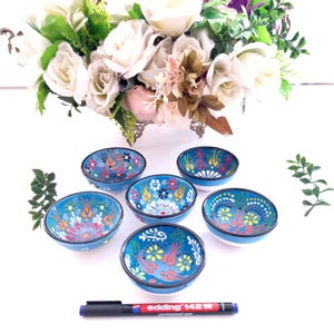 May include: Six small, round, blue ceramic bowls with intricate floral patterns in red, yellow, and white. Each bowl has a black rim and a white base. A black marker with red text is at the bottom. A floral arrangement is in the background.