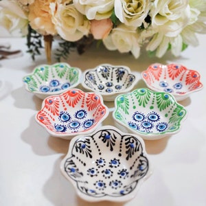 Puede incluir: Seis cuencos pequeños de cerámica blanca con bordes festoneados. Cada cuenco está decorado con un patrón diferente de diseños de ojos malos azules, verdes y rojos.