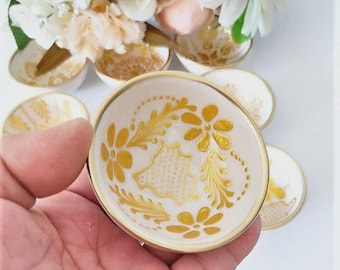 May include: A small, round, white ceramic bowl featuring a gold-painted floral pattern and a gold-trimmed rim. The bowl is held in a palm, with several identical bowls arranged in the background, each decorated with matching gold floral designs.