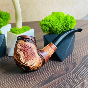 May include: A carved wooden tobacco pipe with a black stem. The bowl features a detailed relief of a wolf's head. The pipe is made of light and dark brown wood, with a gold-colored band near the stem.