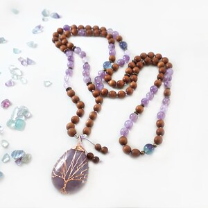 May include: A purple amethyst mala necklace with a tree of life pendant, made with sandalwood beads and fluorite accents.