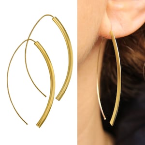 May include: A pair of gold hoop earrings with a curved, slightly flattened shape. The earrings are shown on a model's ear.
