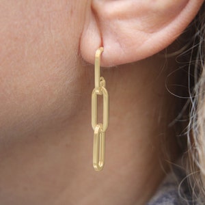 May include: Gold chain earrings with three oval links. The earrings are dangling from a person's ear.