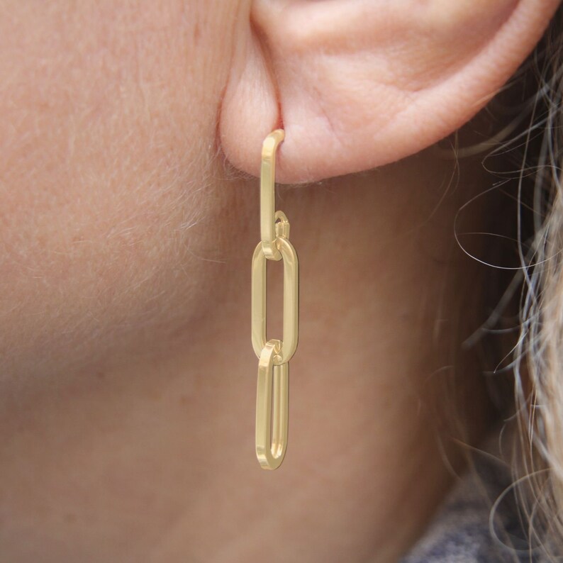 May include: Gold chain earrings with three oval links. The earrings are dangling from a person's ear.