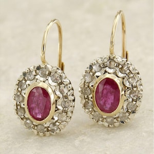 Peut inclure: Une paire de boucles d'oreilles dorées avec des pierres précieuses ovales rouge foncé, entourées de petites pierres claires. Les boucles d'oreilles ont une fermeture à levier.