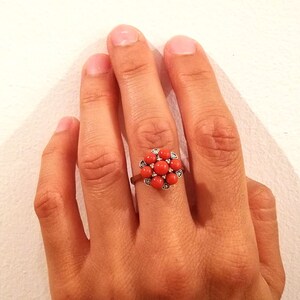 May include: A silver ring with a red coral flower design. The flower has a cluster of small, round coral stones surrounded by smaller, clear stones.