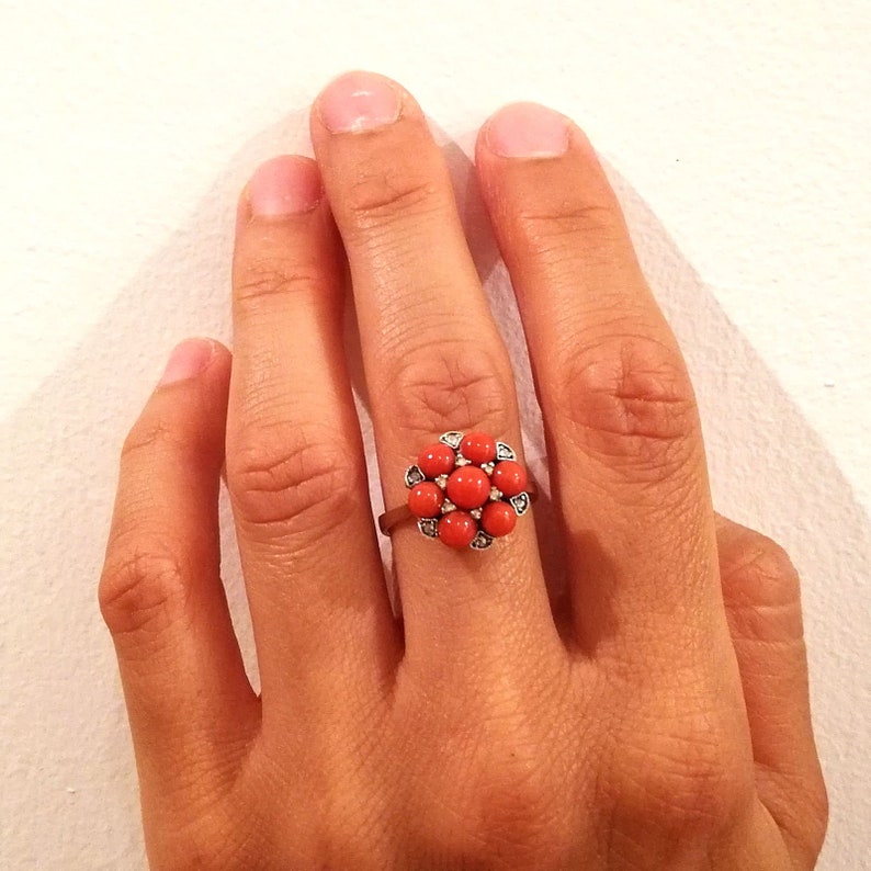 May include: A silver ring with a red coral flower design. The flower has a cluster of small, round coral stones surrounded by smaller, clear stones.