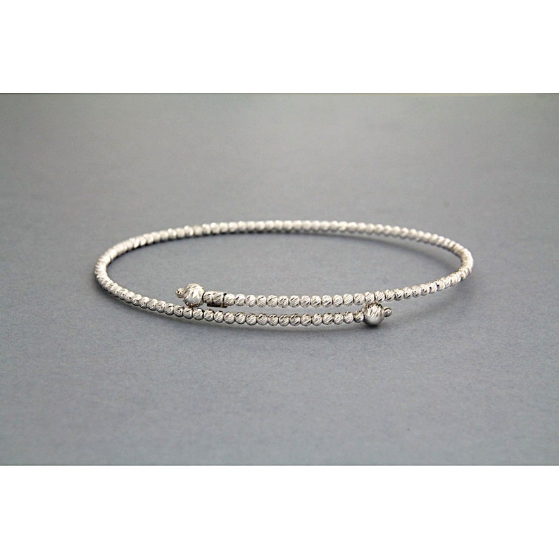 May include: A silver bangle bracelet with a textured finish and two small round beads at the clasp.