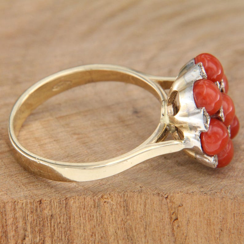 May include: A gold ring with a cluster of red coral stones set in a white metal setting. The ring is on a wooden surface.
