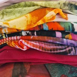 May include: A stack of colorful fabric scraps in various patterns and textures, including floral, striped, and abstract designs. The fabrics are arranged in a neat pile, with the top layer being a light pink fabric.