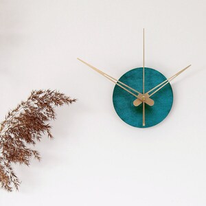 Könnte beinhalten: Eine runde tealblaue Uhr mit goldenen Zeigern an einer weißen Wand, eine hölzerne Gelenkpuppe, ein Stapel Bücher und ein goldenes geometrisches Objekt auf einer weißen Kommode mit vier Schubladen.