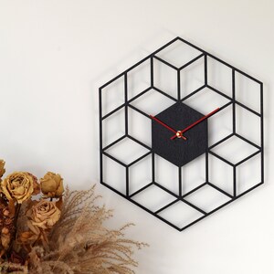 May include: A black geometric wall clock with red hands. The clock is in the shape of a hexagon and has a 3D effect. The clock is hanging on a white wall.
