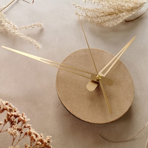 Op de afbeelding: Een gouden klok met een beige wijzerplaat en gouden wijzers. De klok staat op een beige achtergrond met gedroogde bloemen.