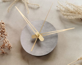 Puede incluir: Un reloj redondo gris con agujas doradas. El reloj está sobre un fondo beige claro.
