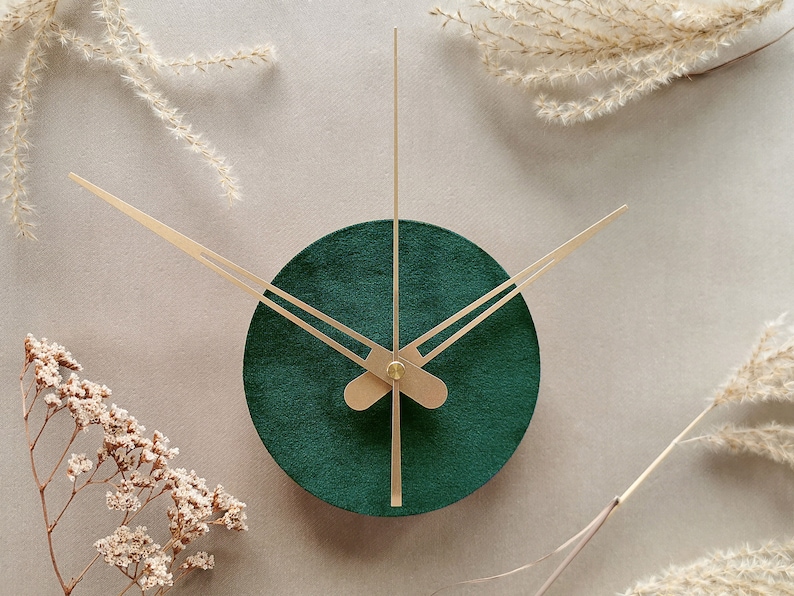 May include: A round, green clock with gold hands. The clock is set against a light gray background with dried flowers around the edges.