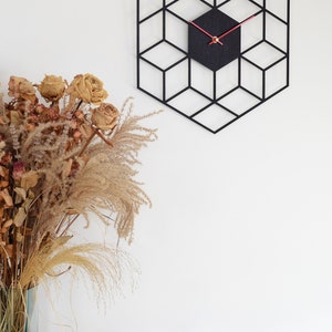 May include: Black geometric wall clock with red hands and a black background. The clock is hanging on a white wall with a bouquet of dried flowers and decorative balls on a shelf below.