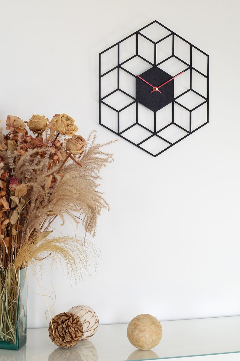 May include: Black geometric wall clock with red hands and a black background. The clock is hanging on a white wall with a bouquet of dried flowers and decorative balls on a shelf below.