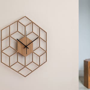 Peut inclure: Une horloge en bois avec un design géométrique. Le cadran de l'horloge est un hexagone avec des aiguilles noires et un fond en bois. L'horloge est montée sur un mur blanc.