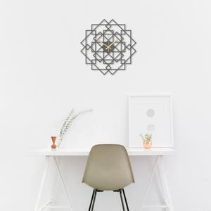 May include: A white desk with a black chair and a geometric clock on the wall. The desk has a white frame and a white top. The chair is a simple black chair with a white seat. The clock is a black metal clock with a geometric design. The clock is hanging on a white wall.