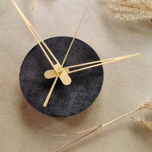May include: A black clock with gold hands on a beige background. The clock face is round and has a textured surface.